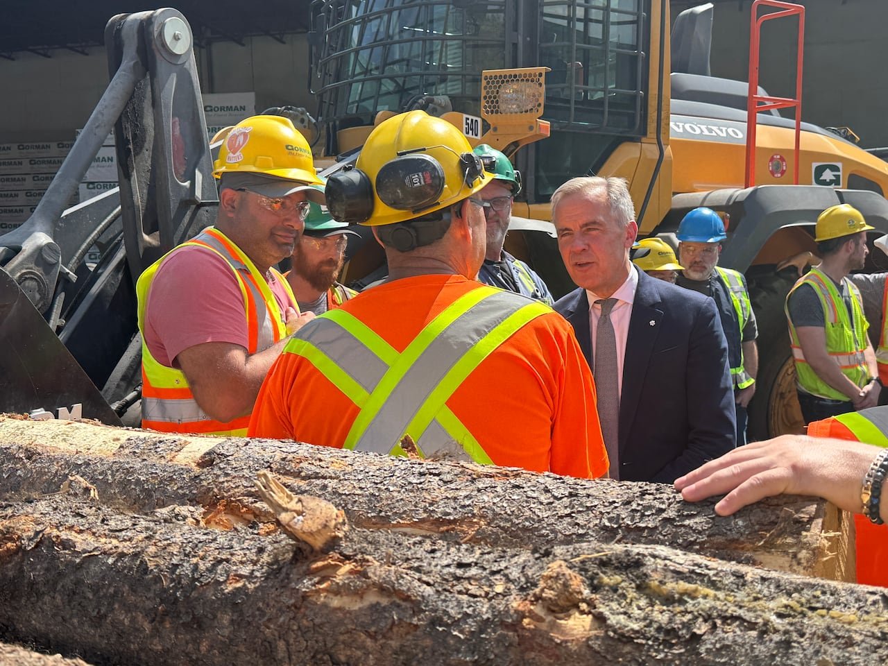 A man in a suit speaks to mill workers in hard hats with logs surrounding them